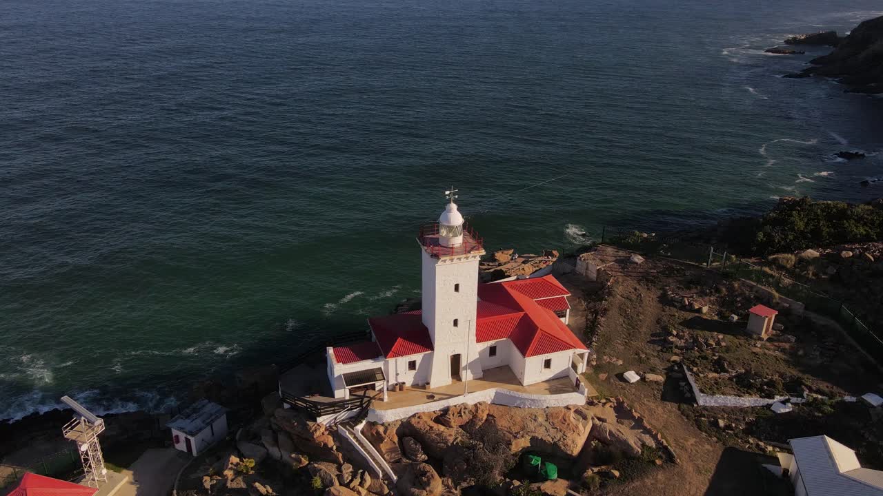 A scenic aerial view of a white lighthouse with a red roof perched on a rocky coastline beside the vast ocean.
