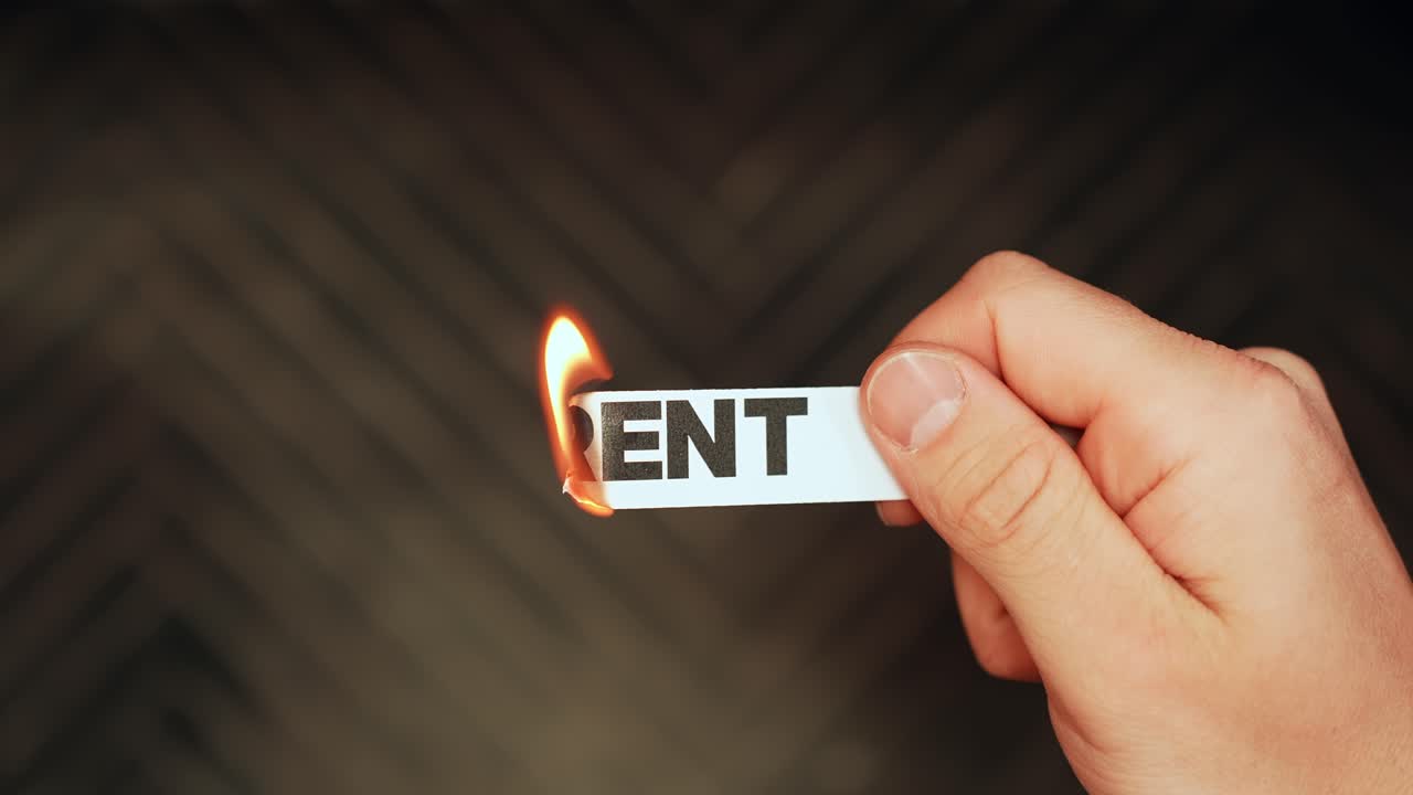 Close-up of a hand holding a paper strip labeled “RENT” as the edge burns with flame on a dark background. Housing costs and affordability concept with dramatic copy space