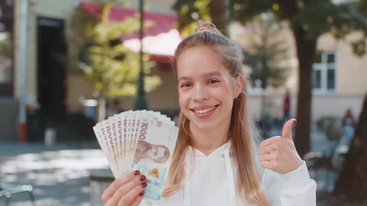 Happy caucasian young woman girl holding fan of euro showing thumbs up gesture on city street