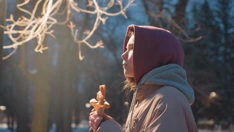 side view of woman in winter coat walking through park and eating snack wrapped in paper, enjoying food outdoors in urban setting with street lights in background