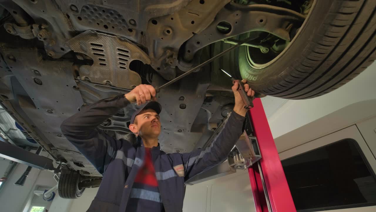 joven mecánico de automóviles en una estación de servicio de reparación inspeccionando la rueda del automóvil y el detalle de la suspensión de un automóvil levantado. vista de abajo.