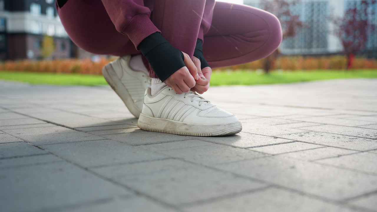 Low angle shot of female jogger bending down on paved sidewalk tying sneaker lace in urban park near modern apartments wearing maroon pants and hoodie before morning run