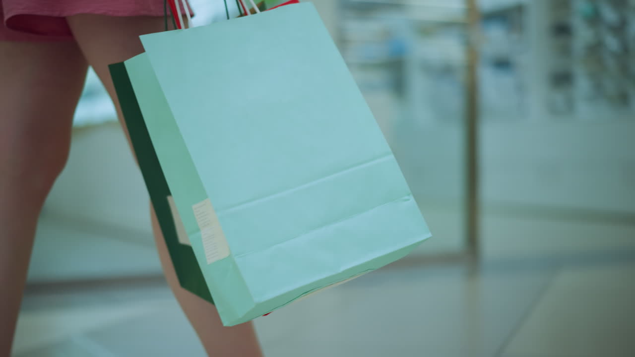 vista de la pierna de la dama en vestido rosa caminando con una bolsa de compras verde y roja de menta en un centro comercial brillantemente iluminado, el fondo muestra artículos de la tienda borrosos, mientras se mueve con calma a través del piso