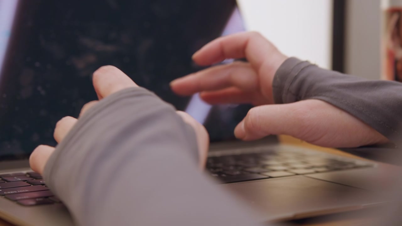 Primer plano de manos en el touchpad del portátil con iluminación tenue y cálida, mostrando mangas de guantes sin dedos, movimiento lento y deliberado del cursor y toques suaves, escritorio acogedor con taza desenfocada, escritor creativo editando texto.