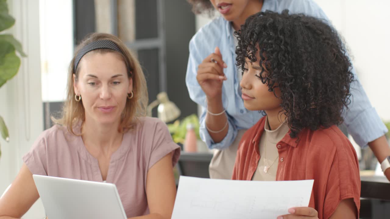Diverse female team meeting at office as standing coworker pointing pen at laptop checking paper