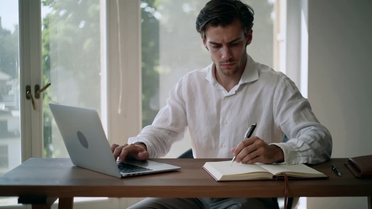 Mann i hvit skjorte leser laptopskjerm, taster og skriver notater hjemme med bærbar PC og notatbok.