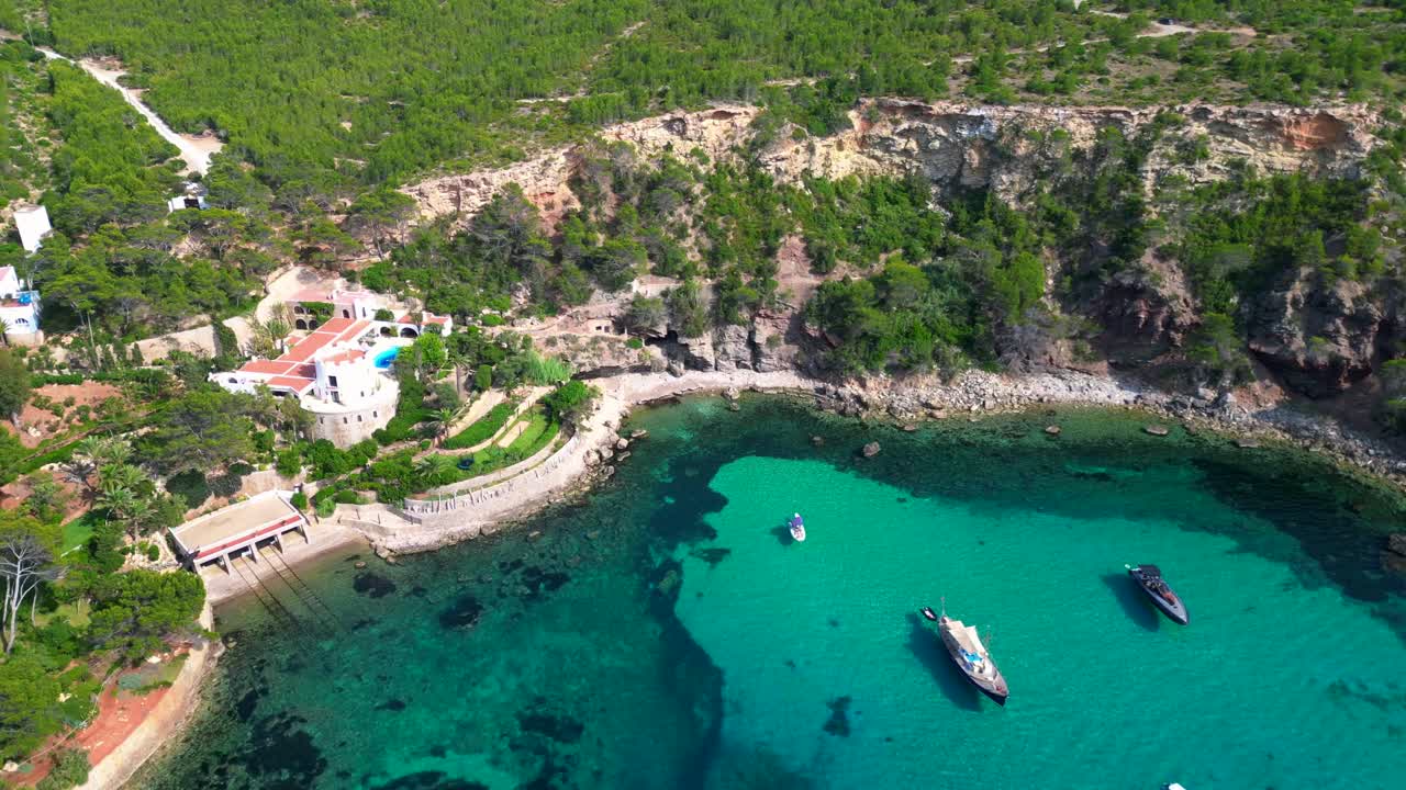 luxury villa in Ibiza, Spain, overlooking a picturesque bay with turquoise water and boats sailing on a sunny summer day. Smooth aerial view flight panorama orbit drone