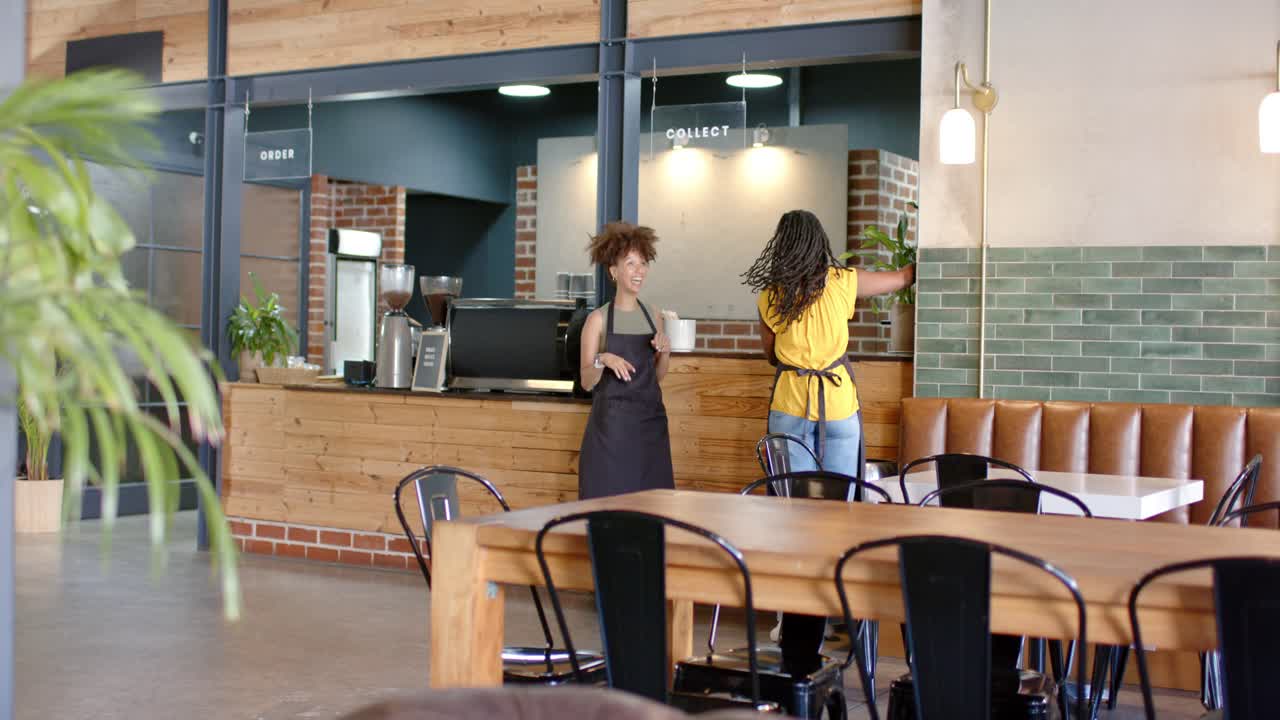 Donne dello staff in grembiule ispezionano le luci indicando il muro, con un cartello "ORDINA" al bancone del caffè con un telefono.