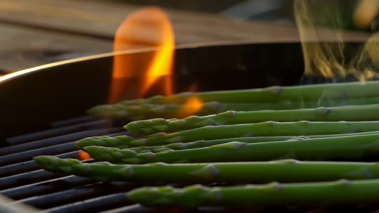Fresh green asparagus spears rest diagonally on dark grill grates as steam, thin smoke, and small orange flames rise around them, creating warm outdoor cooking scene with glossy texture and crisp
