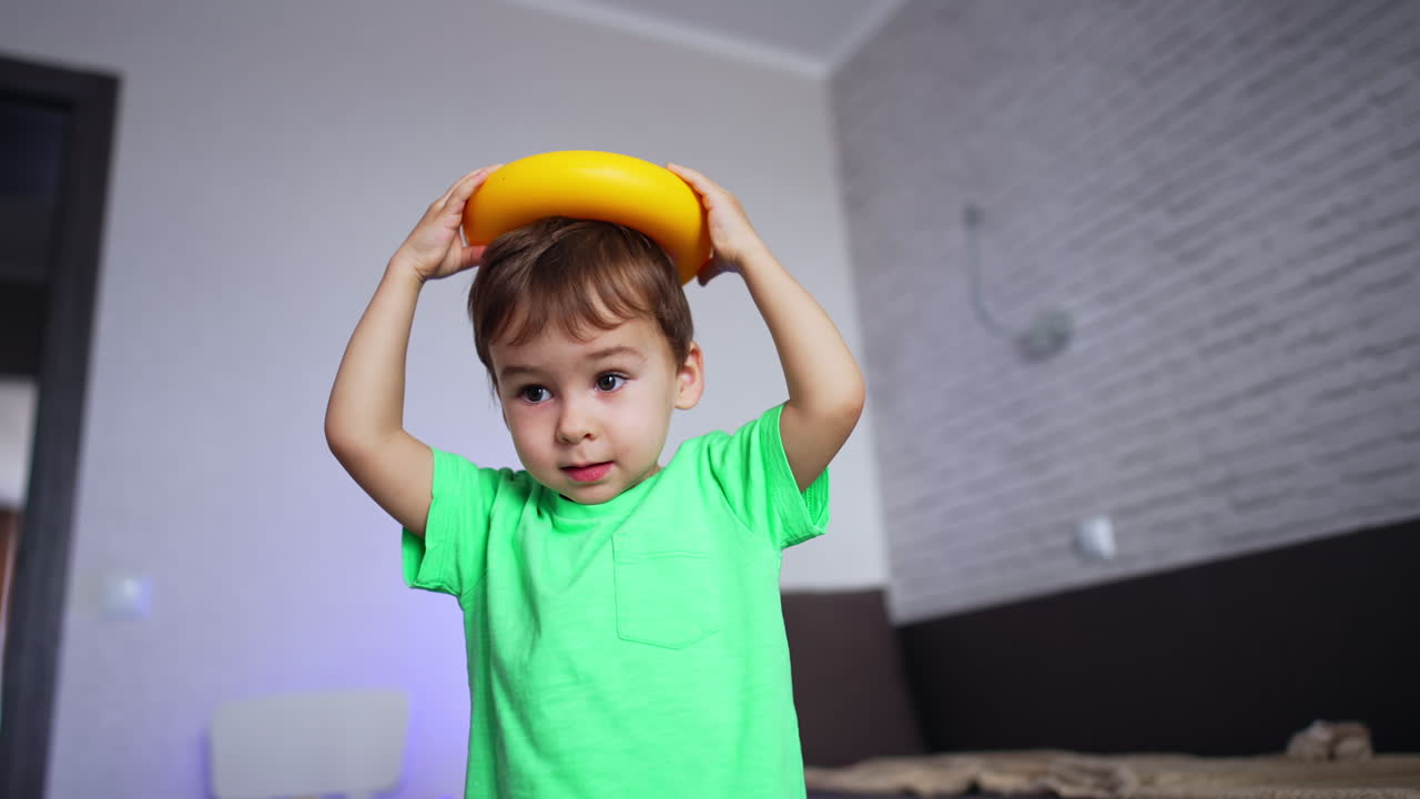 Beautiful Caucasian toddler plays with yellow ring from toy pyramid. Baby boy puts a toy on the head. Low angle view.