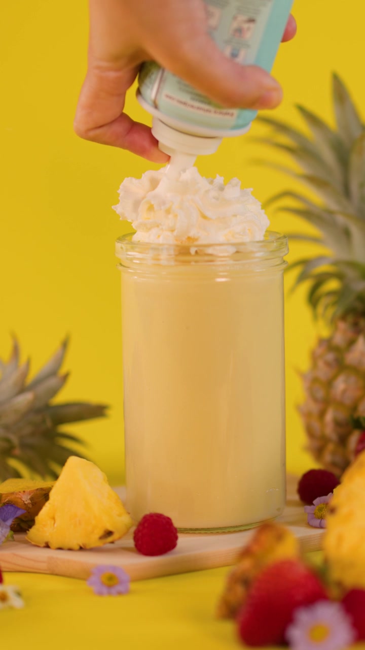 Una mano aggiunge panna montata a un frullato all'ananas con frutta fresca su uno sfondo giallo vivace.