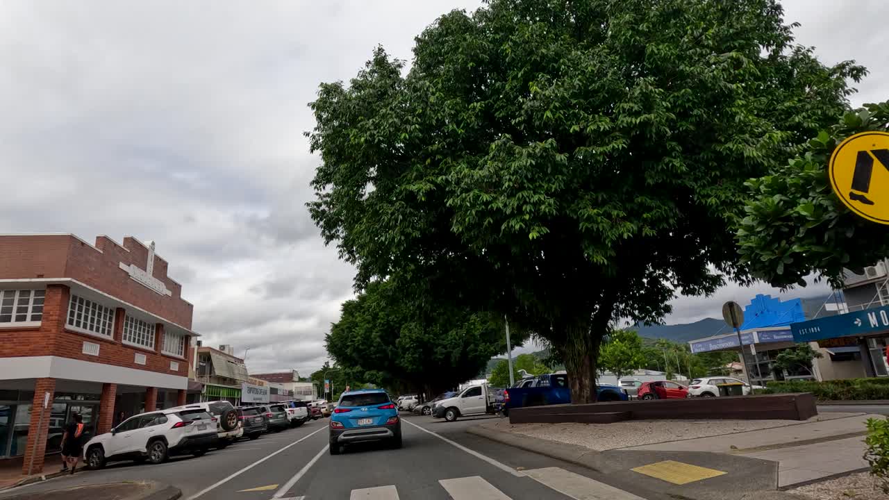 Vehicle drives down urban street lined with shops, palm trees, parked cars, under overcast skies