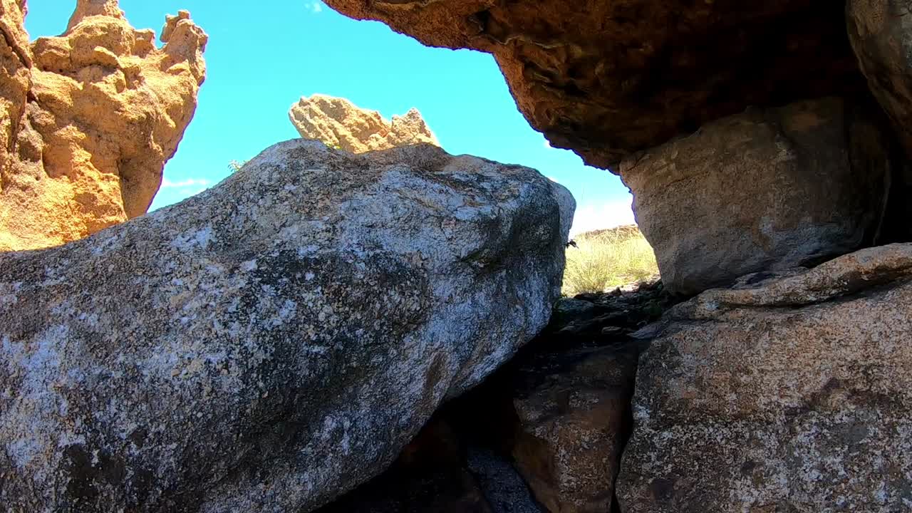 Underneath a large boulder making a natural cave with a large rock formation in the background.