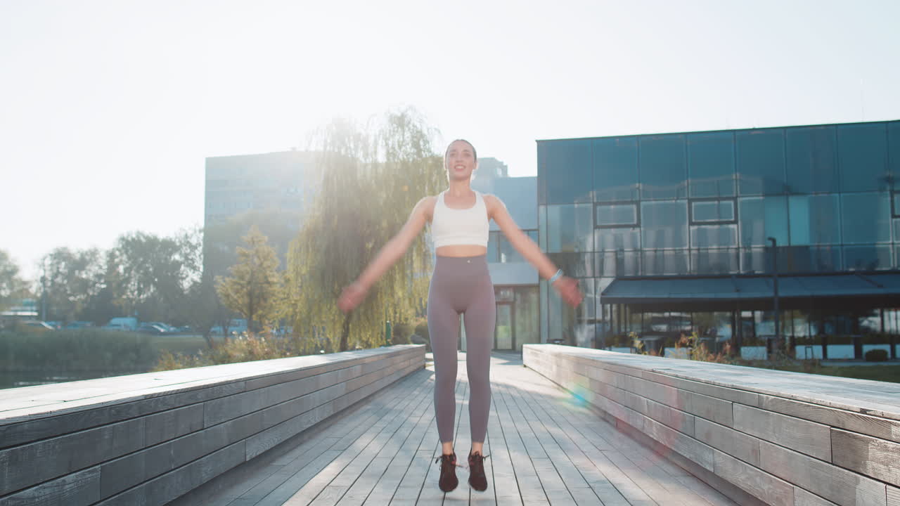 Caucasian sporty young woman jumping on bridge warmup before jogging in city park on sunny day