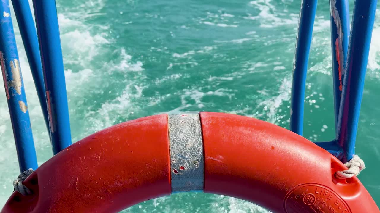 Close-up of a red life buoy with blue railings and churning turquoise water in the background.