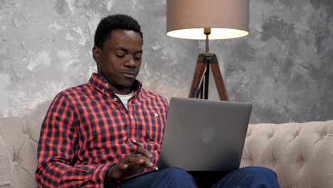 hombre afroamericano sonriente sentado en el sofá trabajando para una computadora portátil escribiendo en el teclado