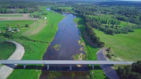 Vista-Aérea-De-Un-Río-Venta-En-Un-Día-Soleado-De-Verano,-Exuberantes-árboles-Y-Prados-Verdes,-Hermoso-Paisaje-Rural,-Disparo-De-Drones-De-Gran-Angular-Que-Avanza-Sobre-El-Puente-De-Hormigón-Blanco