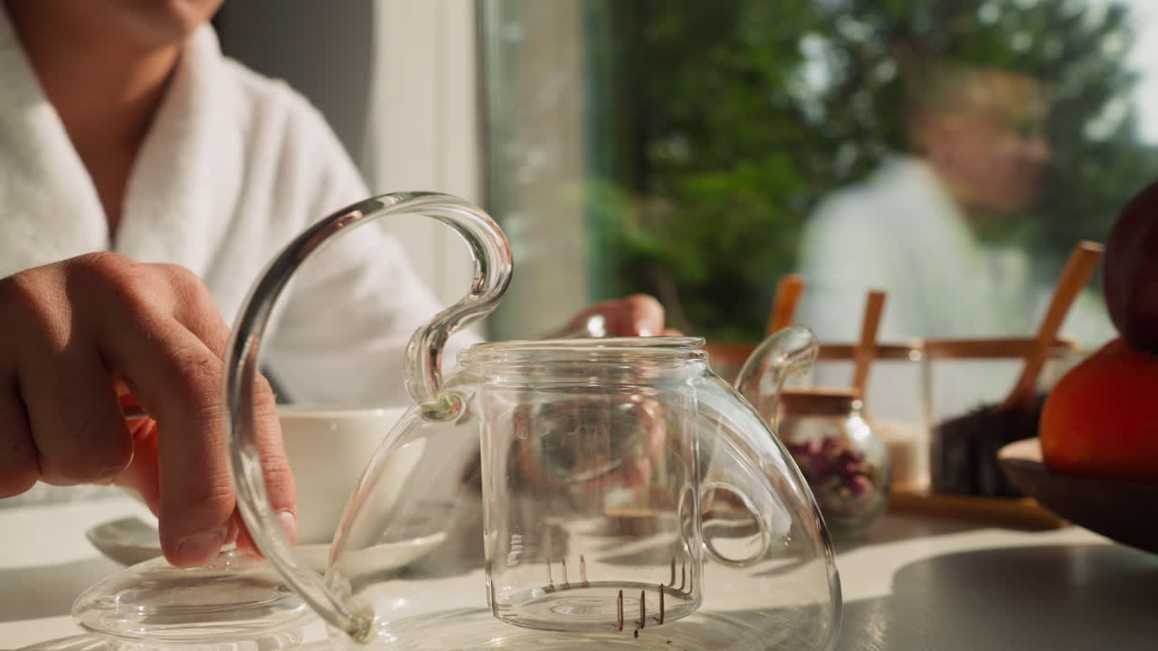 el hombre abre la tapa de una elegante tetera de vidrio para preparar té. el joven disfruta de la ceremonia del té todas las mañanas. el hombre prefiere la mezcla de té natural para la bebida matutina