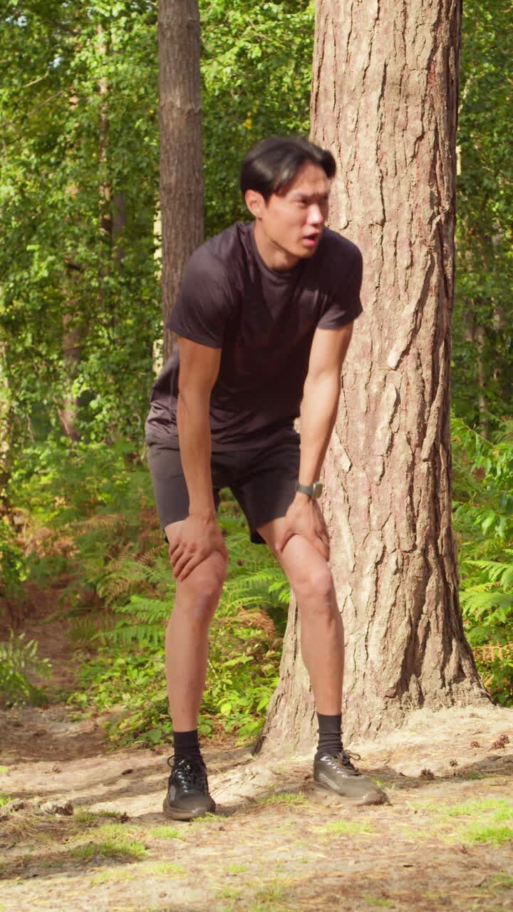 Video verticale di un uomo che si allena all'aperto, correndo lungo un sentiero nella foresta, fermandosi per riprendere fiato e controllando l'app di attività sullo smartwatch, ripreso in tempo reale