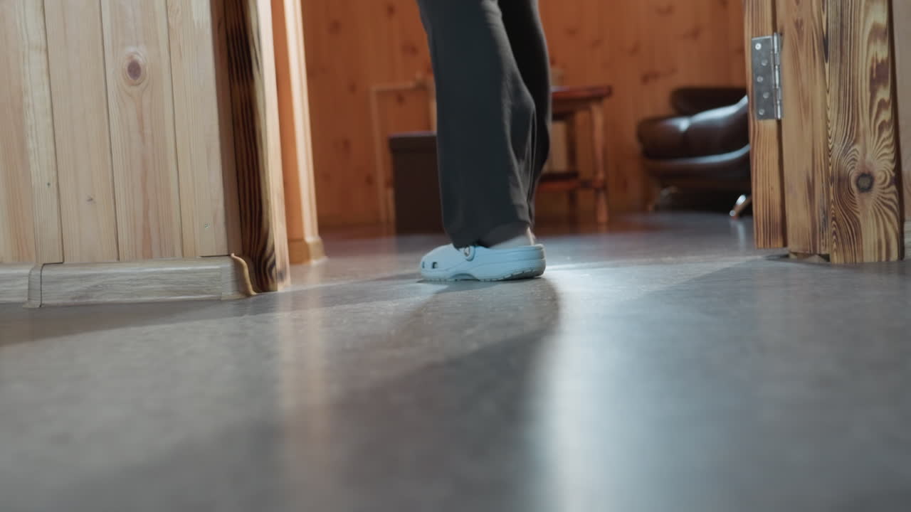 low angle leg view of individuals walking out of wooden room wearing casual slippers and denim trousers, casting long shadows on floor in cozy interior with warm lighting and relaxed atmosphere
