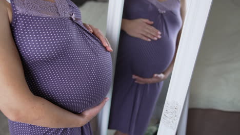 woman on the last months of pregnancy standing in front of the mirror. pregnant lady in violet sleepwear watching at her reflection in the mirror.
