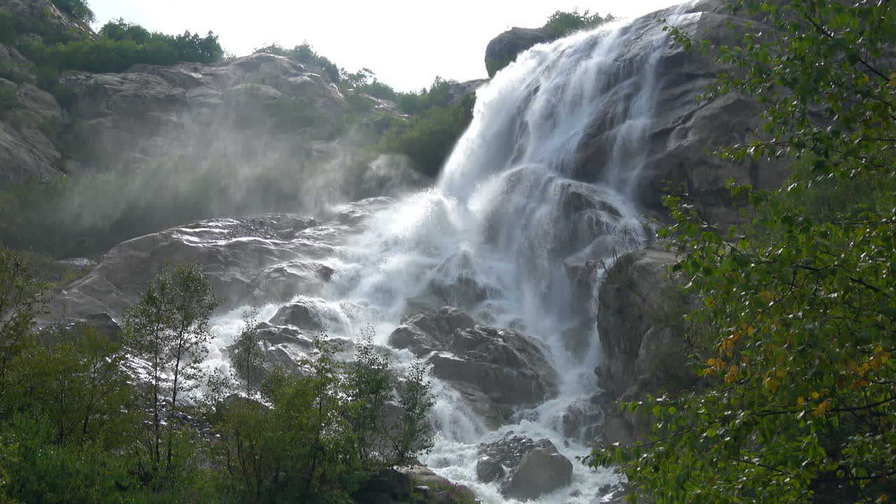 Air terjun pegunungan yang perkasa mengalir deras di atas bebatuan gelap dengan kabut dan hutan hijau di sekitarnya, direkam di Dombay, Kaukasus, Rusia, pemandangan alam liar musim panas yang indah.