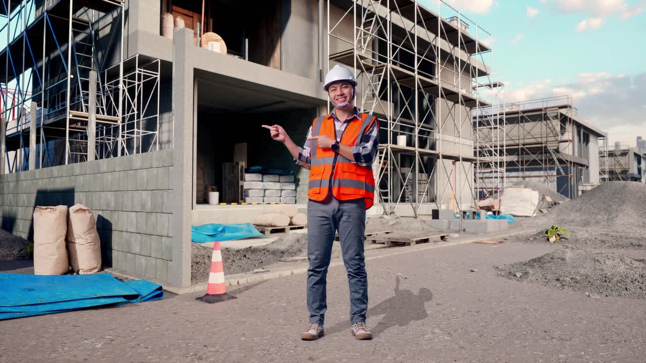 Full Body Of Asian Male Engineer With Safety Helmet Smiling And Pointing To Side While Standing At Building & Construction Site in Progress to New House