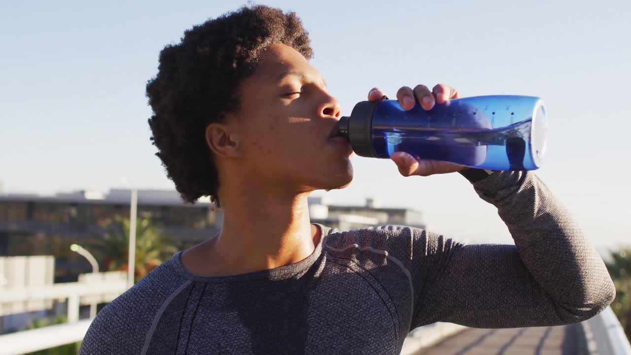 hombre afroamericano en forma haciendo ejercicio al aire libre en la ciudad, descansando, bebiendo de la botella de agua en el sol