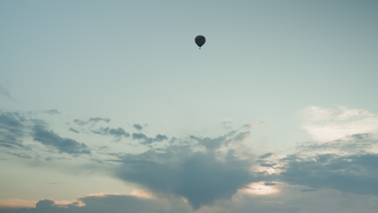 distant view of colorful hot air balloon floating solitary high above expansive green field at dusk with soft pastel sky and drifting clouds evoking sense of adventure freedom