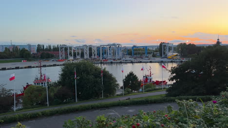 Sunset-view-over-a-marina-with-moored-boats-and-industrial-structures,-flags-fluttering-in-the-wind,-and-a-soft-colored-sky---Westerplatte-Gdańsk,-poland