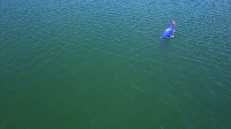 AERIAL: Segelboot im blauen Meer. Ein Segelboot kann am Horizont gesehen werden. Drone view of a single sailboat off the sea. White Sailing Yacht in the Middle of the Boundless Ocean. Aerial View