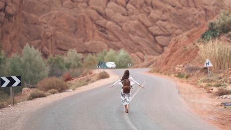 Woman-walking-away-in-the-middle-of-the-road-with-open-arms-at-a-desert-canyon-arid-environment-with-a-rocky-background