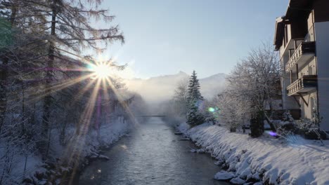 sun shining bright behind snow laden mountains during sunrise, and water stream flowing calmly amidst them