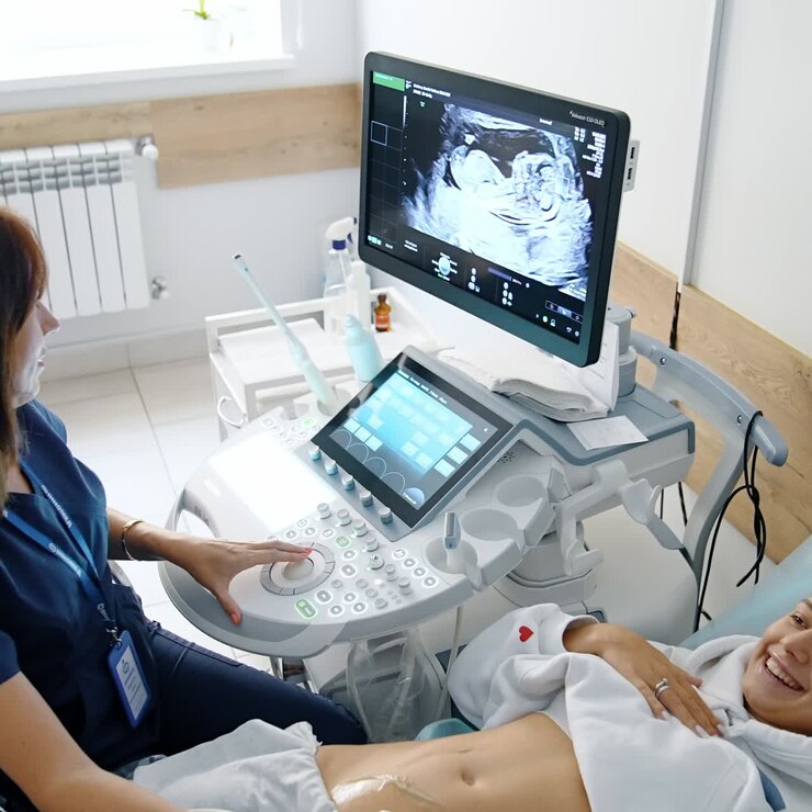 Donna felice ed emozionata distesa sul divano vicino a un'apparecchiatura per ecografie. Bella donna incinta indica lo schermo mentre guarda il suo bambino