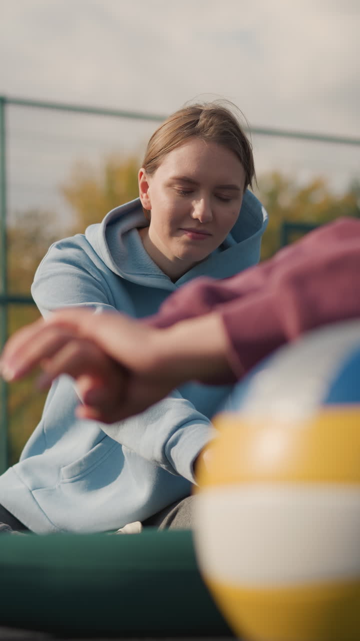 primer plano de tres damas estirándose con una pelota de voleibol cerca, mientras que otras trabajan en el fondo en una arena deportiva al aire libre, mostrando entrenamiento de fitness y trabajo en equipo