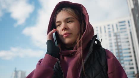 athlete in mauve hoodie and black vest jacket chats on mobile phone with joyful smile under bright sunny sky in urban courtyard framed by modern high rise flats surrounded by leafy greenery