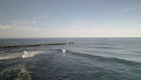 Toma-Aérea-De-Drones-De-Playa-Rápida-Moviéndose-A-Lo-Largo-De-Las-Rocas-Hacia-El-Embarcadero-Y-La-Torre-De-La-Casa-De-Luz-Marcando-El-Canal-Del-Barco