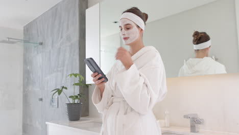 applying face mask, woman in bathrobe using smartphone in bathroom spa routine, at home