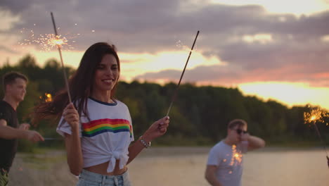 the female student with nude waist is dancing with big bengal lights in her hands on the sand beach with her friends. this is theamy summer evening on the open air party.