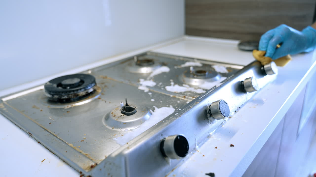 Dirty stovetop surface needing cleaning. Stovetop shows signs of grease and food residue while a gloved hand reaches for a control knob in the kitchen