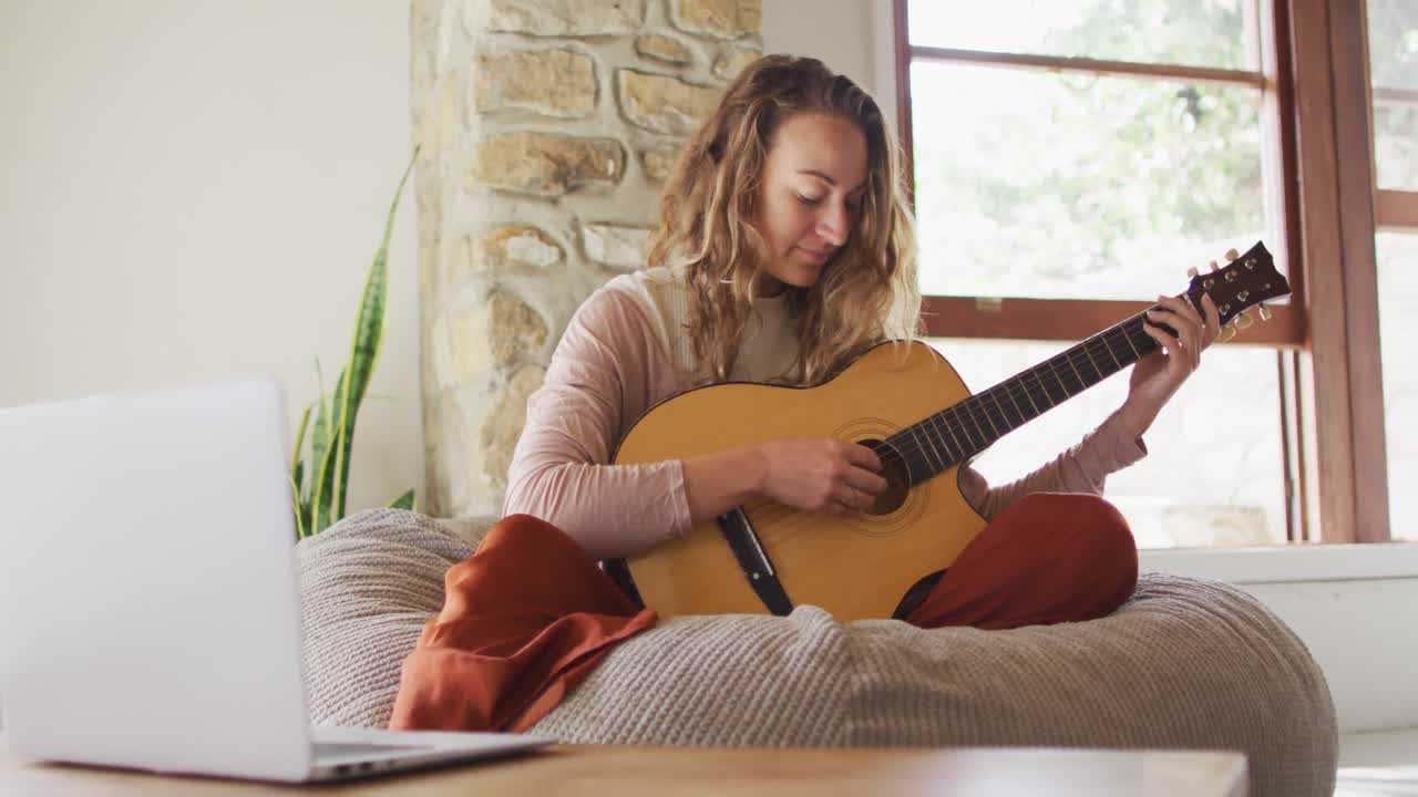 Glad kaukasisk kvinna spelar akustisk gitarr, sittande på en saccosäck i ett soligt vardagsrum i stugan.