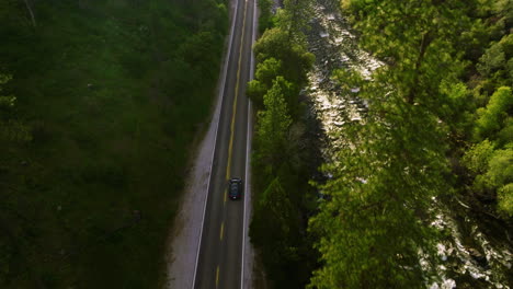 Antena-De-Drones-Siguiendo-Un-Coche-Tesla-Azul-Conduciendo-Por-La-Carretera-Al-Lado-Del-Río-Con-Una-Exuberante-Vegetación-Cerca-Del-Parque-Nacional-De-Yosemite-En-California,-EE.UU.