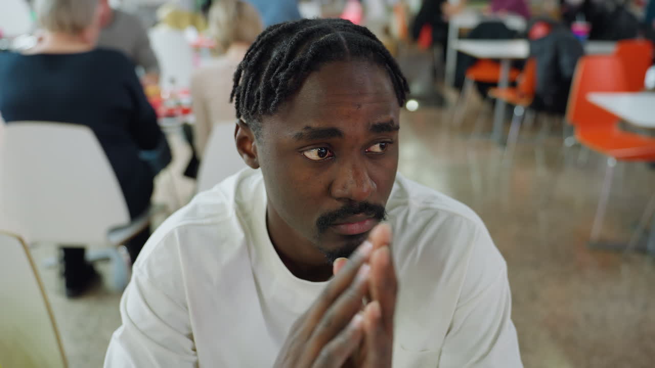 Exhausted young african man wearing white shirt covers both eyes with palms while sitting in busy cafe filled with people showing clear signs of stress fatigue and frustration in public environment