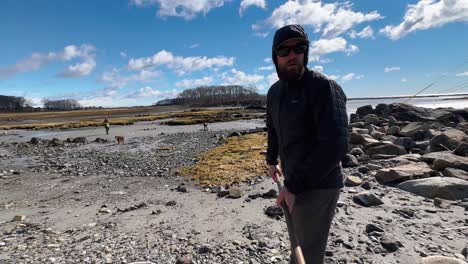 man clears seaweed off of a dark sand beach and plops it into a bucket while a dog is playing fetch in the background