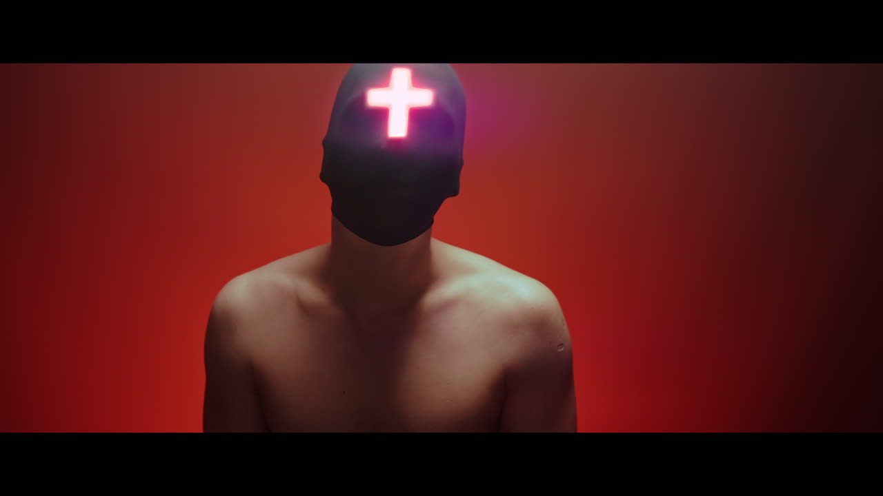 Masked and hooded man with cross of light glowing on his face looks in the camera - red background