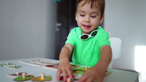 lovely boy in green t-shirt sitting at the desk at home playing with toys. smiling kid takes taps to pieces of puzzles.
