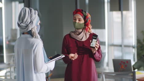 muslim female colleagues in masks using smartphone and speaking