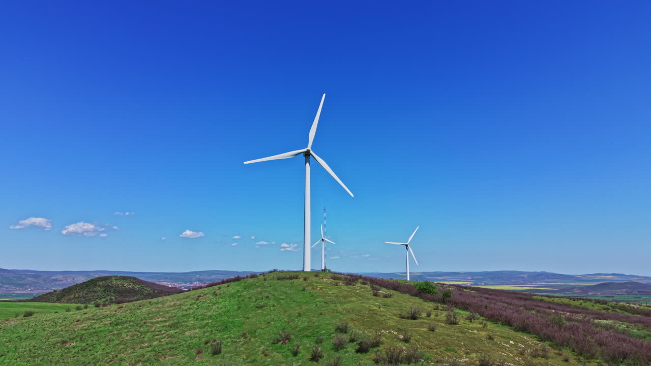 Turbine eoliche si ergono su una collina verde sotto un cielo azzurro e limpido con qualche nuvola in lontananza, catturando energia eolica in una località remota durante il giorno