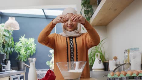 video of smiling biracial woman in hijab baking in kitchen at home, breaking egg into mixing bowl