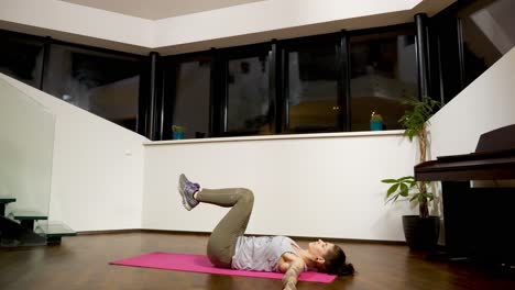 Mujer-Estirándose-Sobre-Una-Estera-De-Yoga-En-El-Interior-De-Una-Casa-Por-La-Noche,-Realizando-Un-Estiramiento-De-Giro-Acostado,-Ambiente-Tranquilo